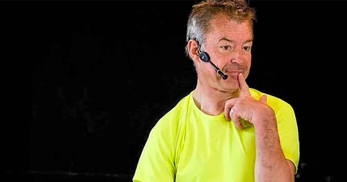 un homme sur scène avec t-shirt jaune fluo, une oreillette et un doigt posé sur la bouche