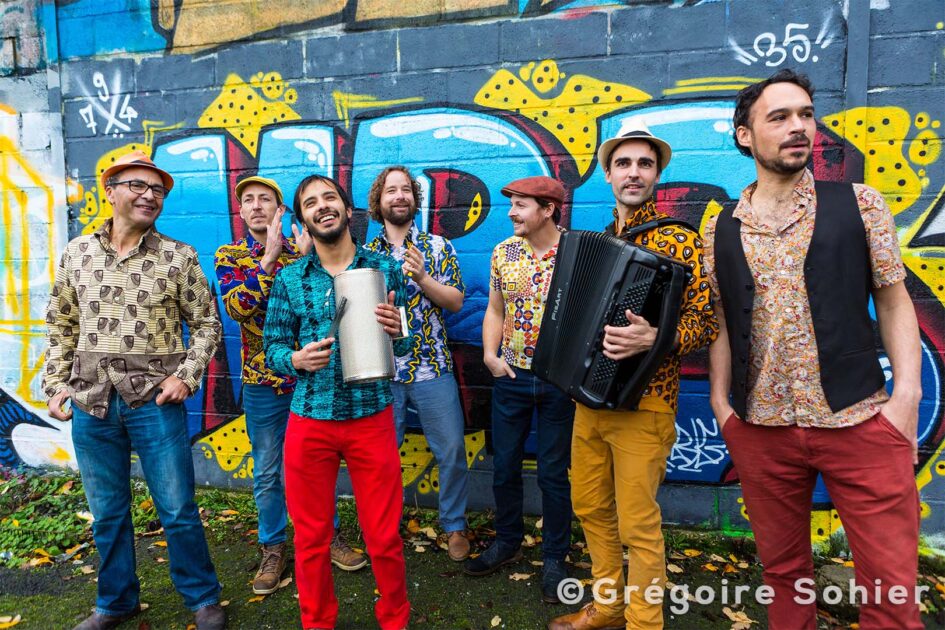groupe de 7 musiciens au vêtements colorés devant un mur avec tags