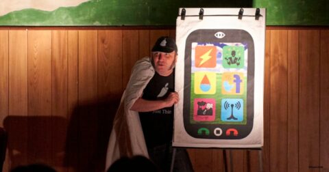 un comédien dans la pénombre devant un paperboard représentant un téléphone portable