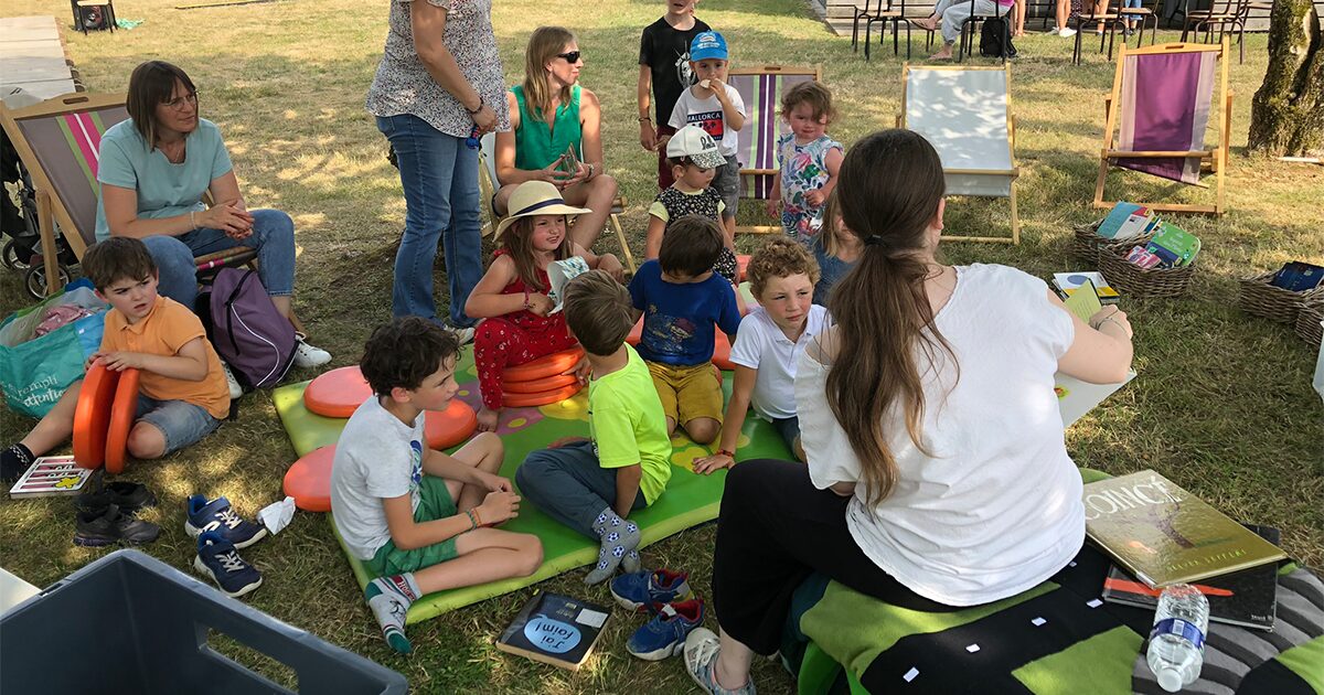 groupe d'enfants et parents lisant des livres dans l'herbe sur des tapis et des transats