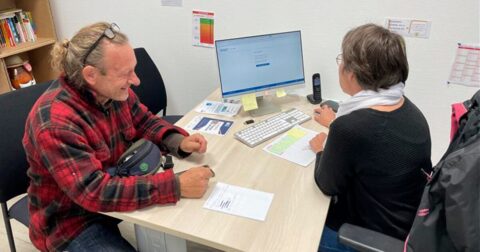 un homme en veste rouge souriant à une femme l'aidant pour ses démarches devant un ordinateur posé sur un bureau