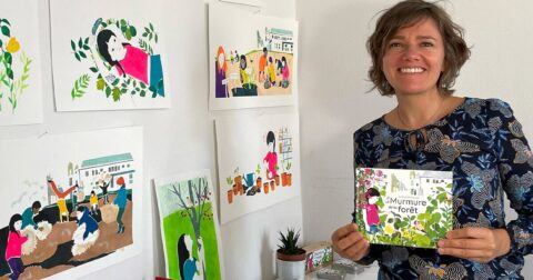 femme souriante tenant un livre devant un mur recouvert de ses dessins