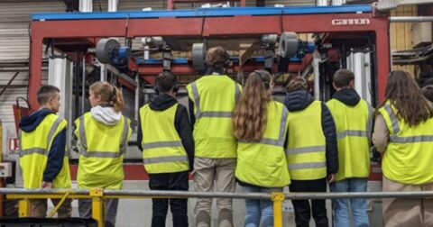Des lycéens portant des gilets jaunes regardent une machine industrielle avec attention.